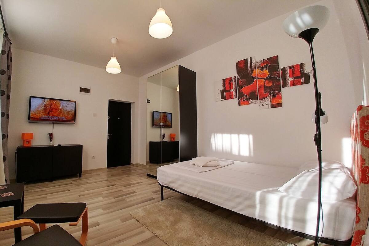 A modern hotel room with a bed, a desk, a TV, and artwork on the wall.