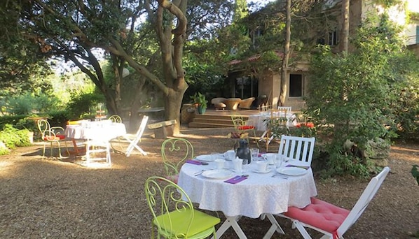 Un espace extérieur aménagé pour les repas, avec des tables et des chaises rondes, entouré d’arbres et avec un bâtiment en arrière-plan.