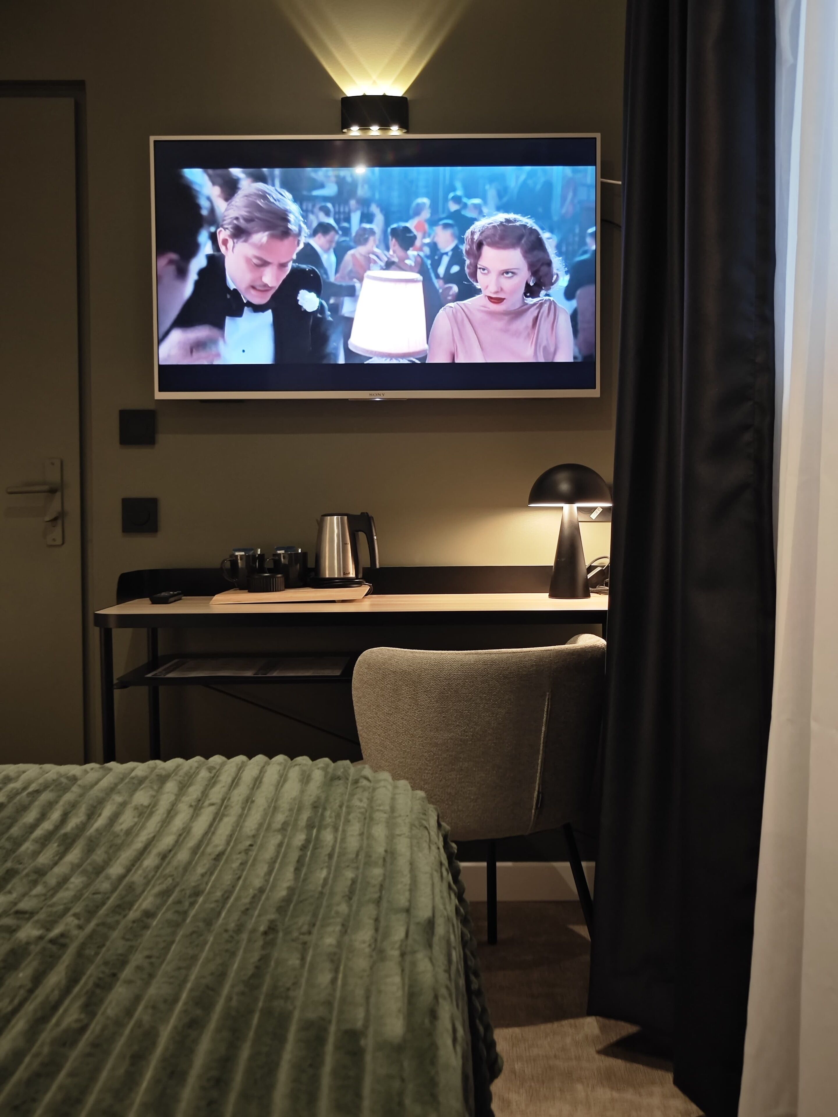 Una habitació d'hotel amb llit, escriptori amb làmpada, cafetera i televisió penjada a la paret.