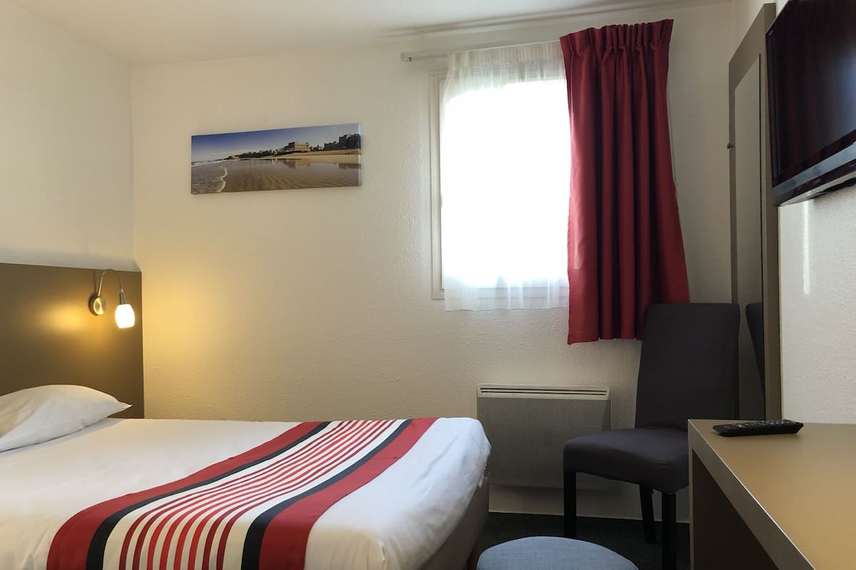A hotel room with a bed, a desk, a chair, a radiator, a window with curtains, and a wall-mounted picture.