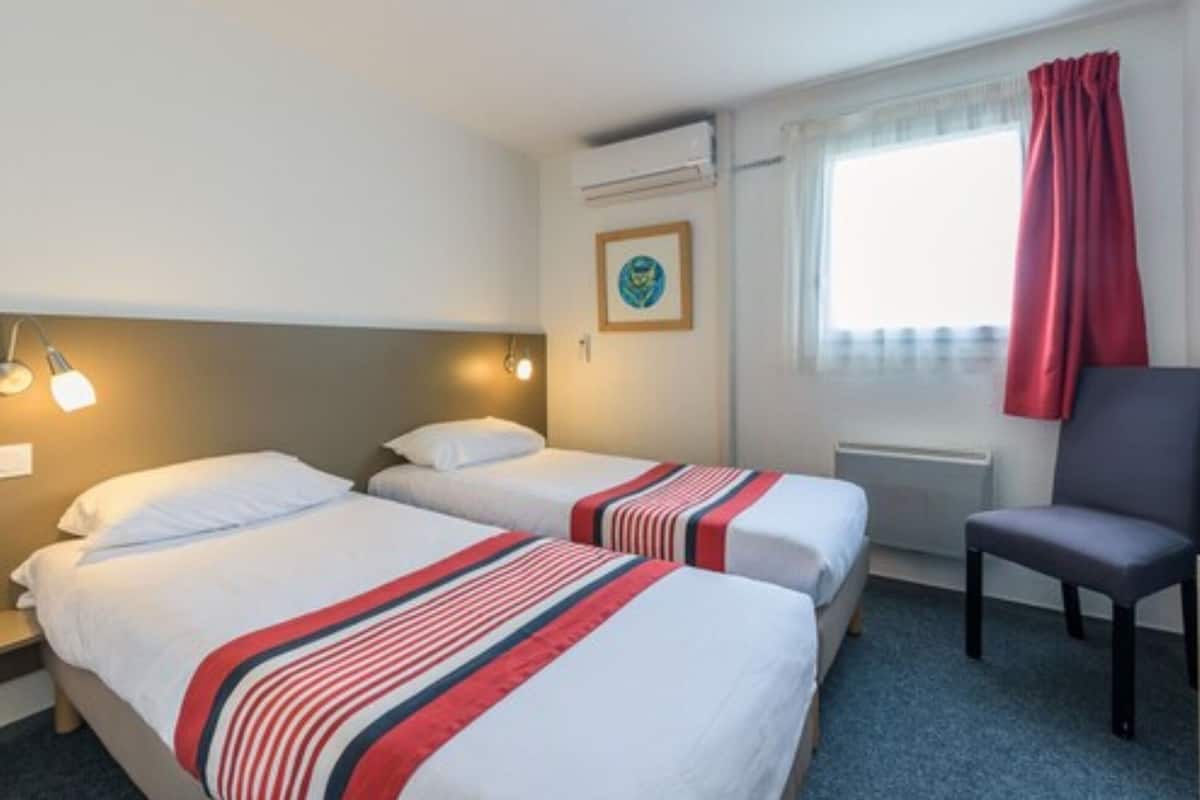 A hotel room with two beds, a chair, a window with curtains, an air conditioning unit, and a framed picture on the wall.