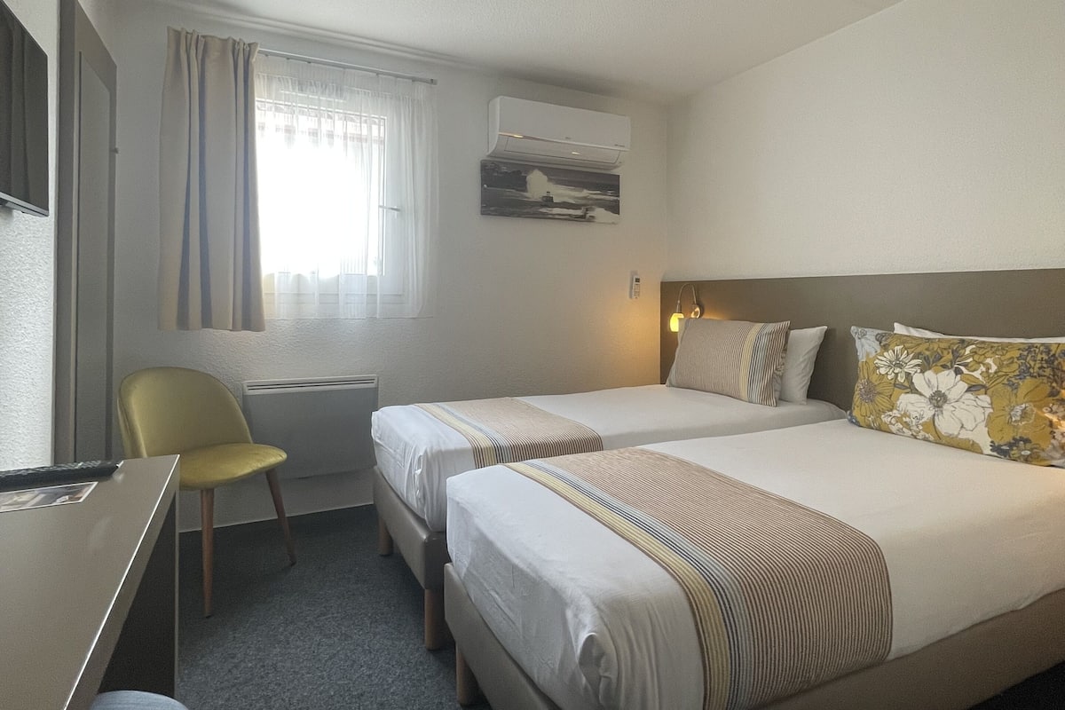 A hotel room with two beds, a desk, a chair, a window with curtains, an air conditioning unit, and a painting on the wall.
