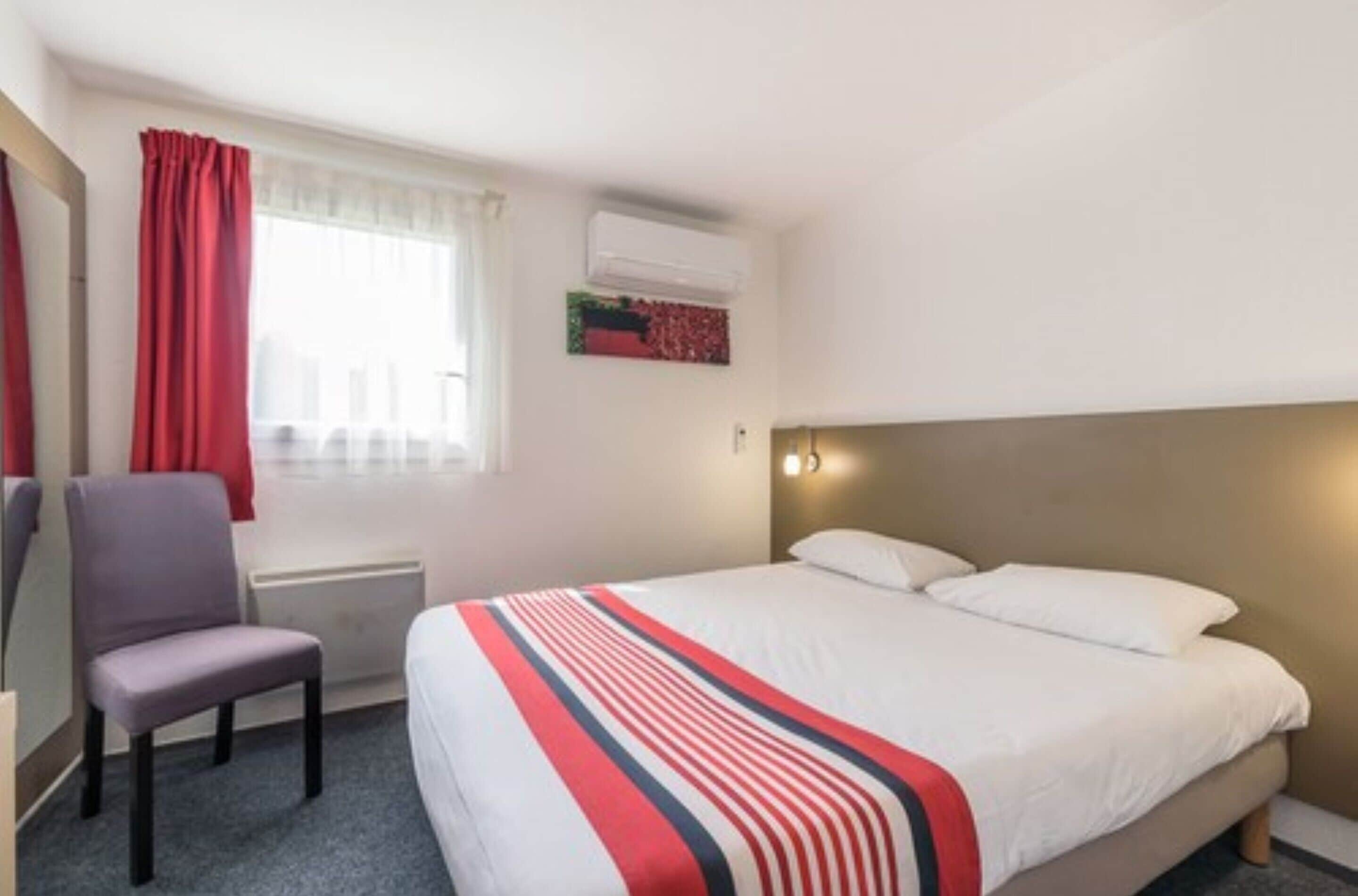 Quarto de hotel com uma cama, uma cadeira, uma janela com cortinas, unidade de ar condicionado e uma obra de arte na parede.