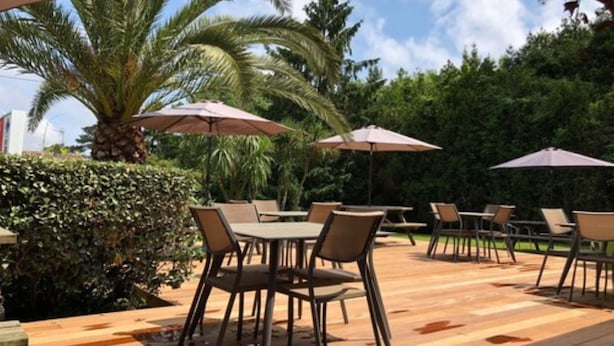 An outdoor patio with wooden flooring, tables, and chairs, surrounded by greenery and palm trees. - Fasthôtel Biarritz Bidart Côte Basque - FH Collection (Bidart)