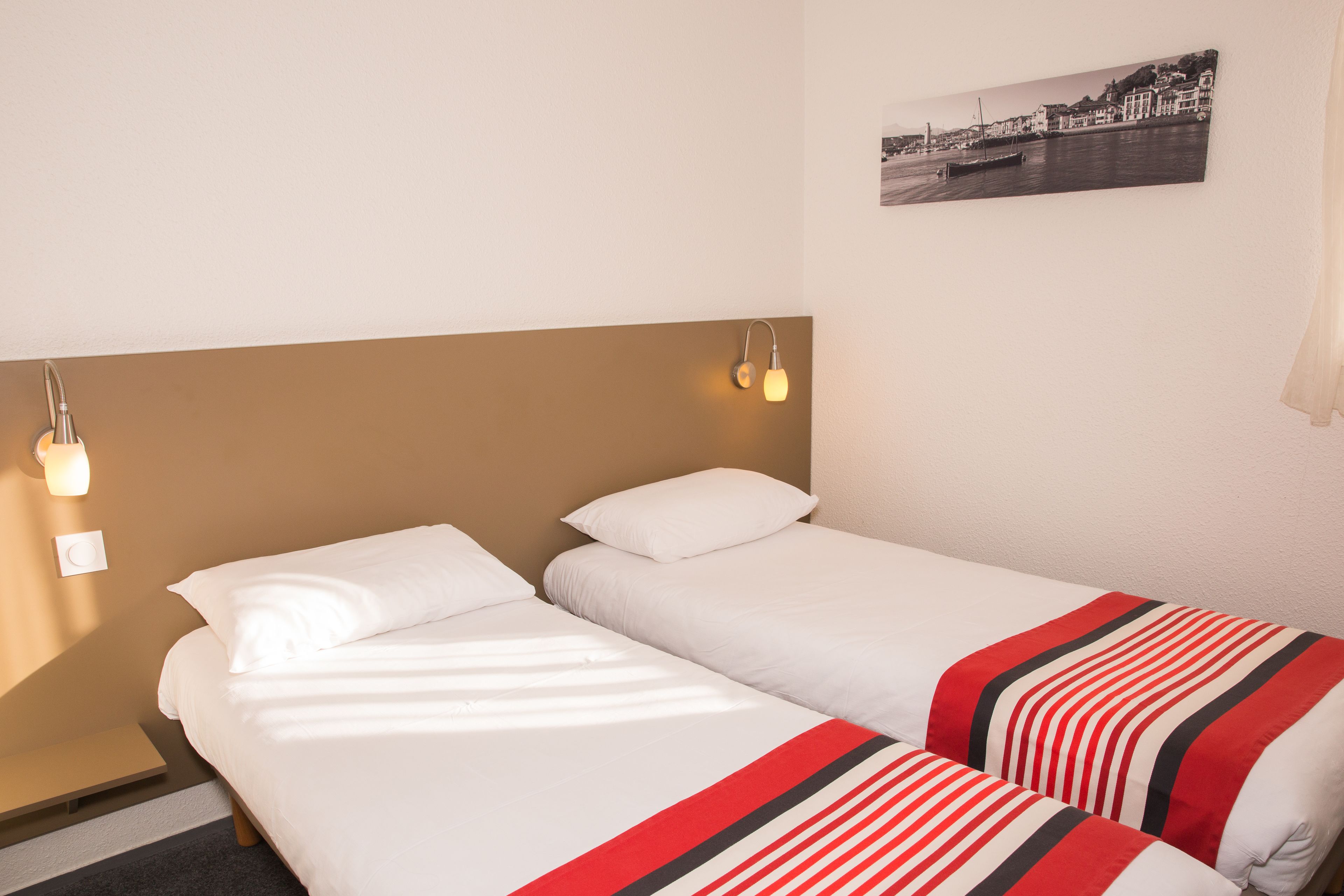 Una habitación de hotel con dos camas individuales, una mesita de noche, una lámpara fijada a la pared y un cuadro enmarcado.