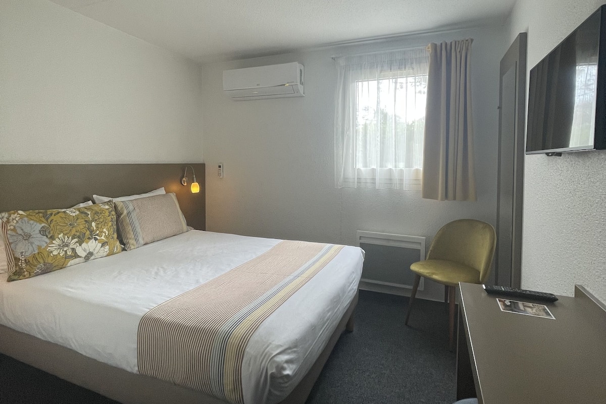 A hotel room with a bed, a desk, a chair, an air conditioner, a window with curtains, and a mirror.