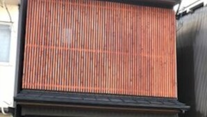 Un edificio moderno con fachada de listones de madera, entrada de color marrón oscuro y suelo de baldosas con una franja de pavimento táctil amarilla.