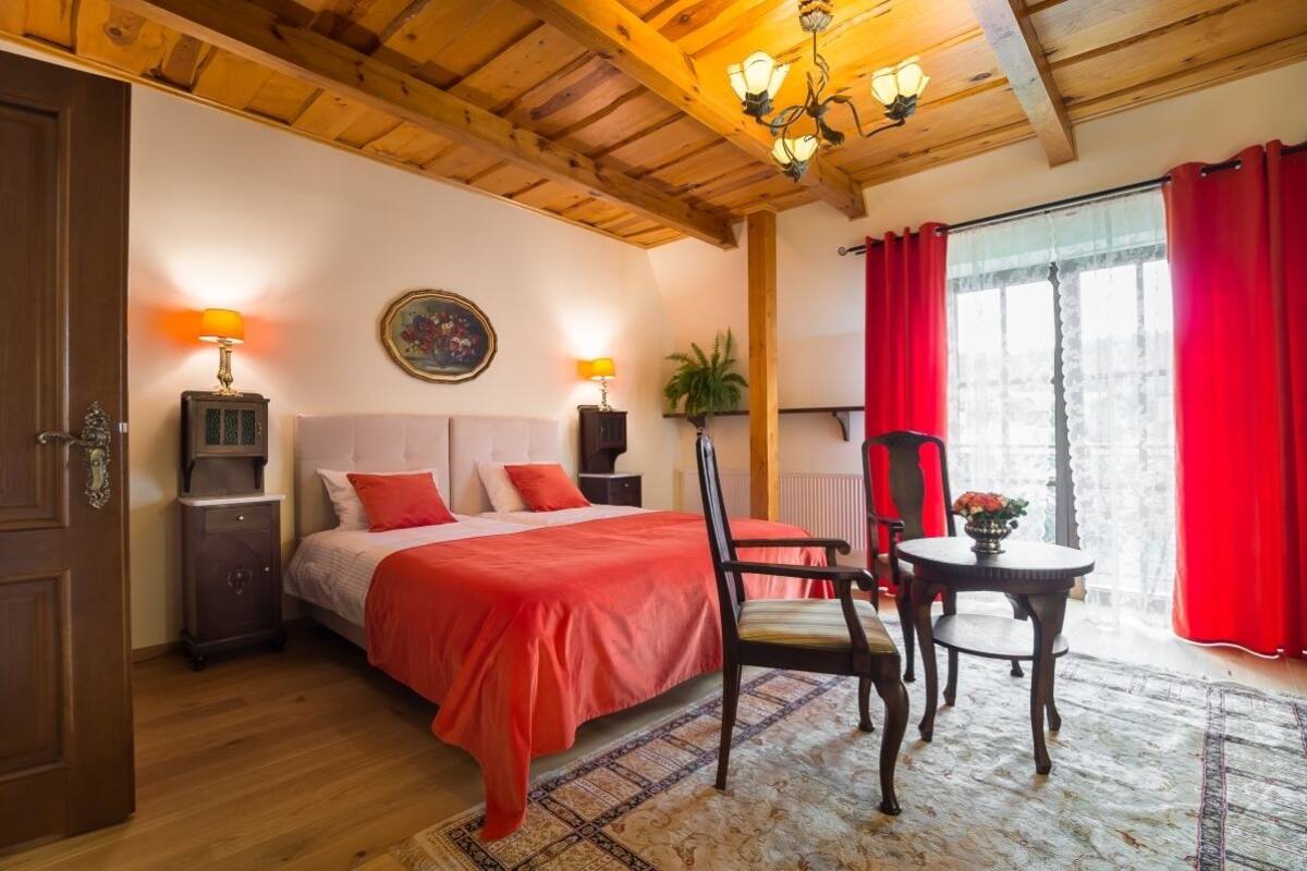 A bedroom with a wooden ceiling, a bed with red bedding, a nightstand, a chair, a table with a vase, and a window with red curtains.