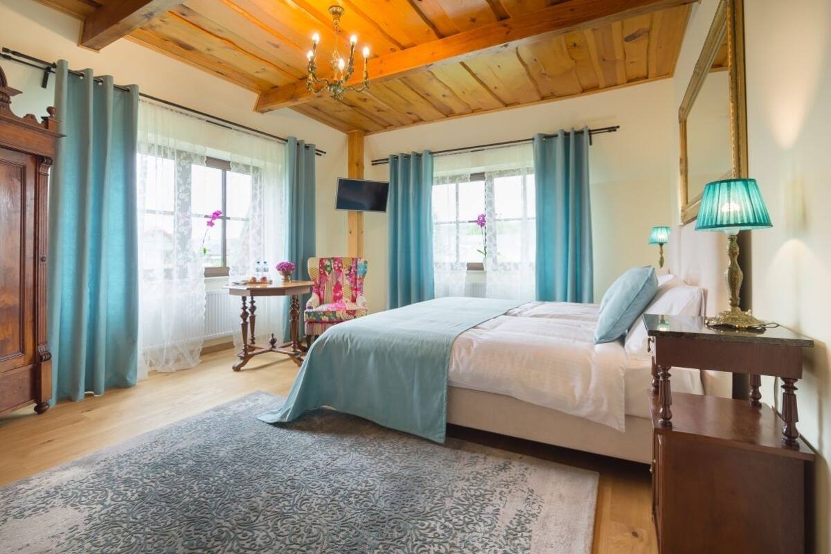 A bedroom with a wooden ceiling, a bed with a blue bedspread, a wooden nightstand, a table with a lamp, and a television on the wall.