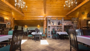 Un intérieur rustique en bois, avec de hauts plafonds, des lustres et une cheminée en pierre. Des tables et des chaises sont disposées pour les repas.