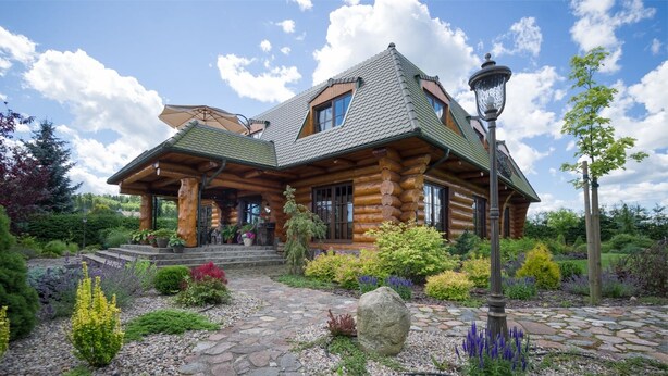 Une cabane en rondins avec un sentier de pierres menant à l’entrée, entourée d’un jardin où poussent différentes plantes et agrémentée d’un grand rocher servant d’élément central.