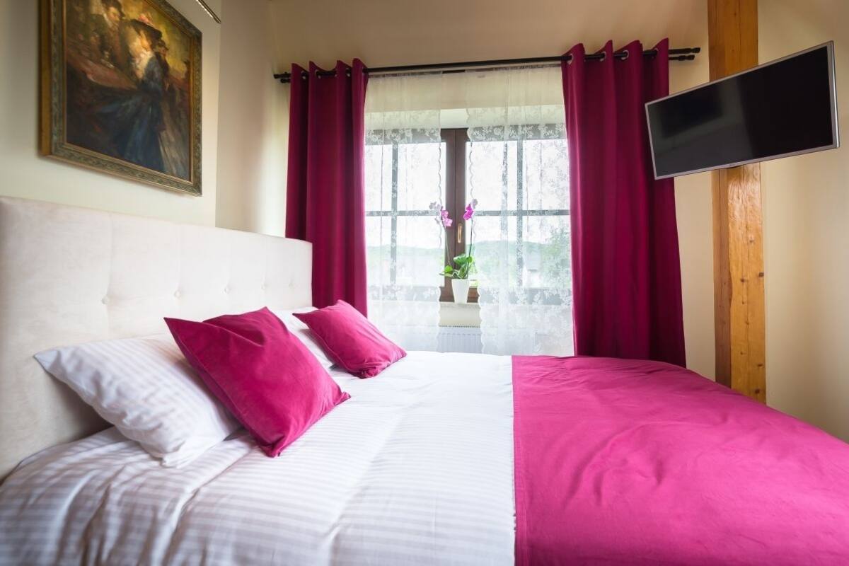 A hotel room with a large bed, red curtains, a flat-screen TV, and a painting on the wall.