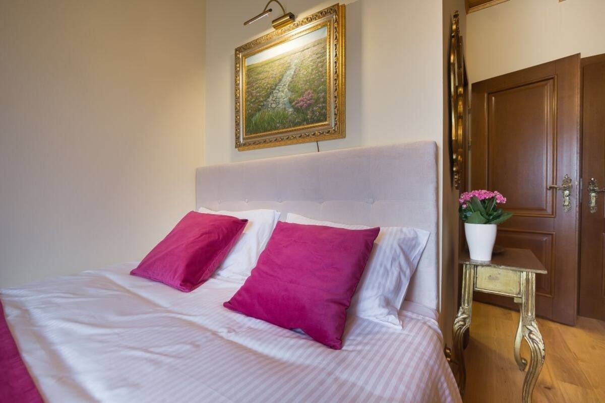 A neatly made bed with white and pink bedding, a framed painting above it, and a wooden door to the right.