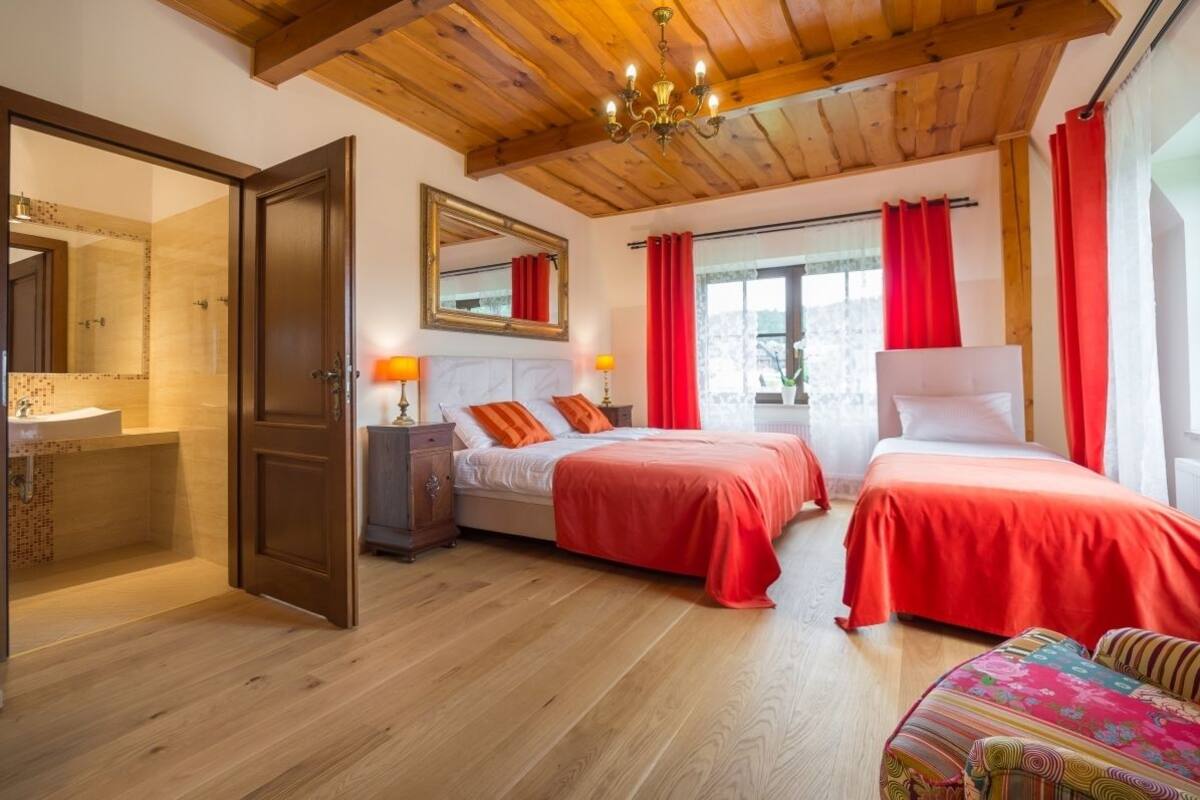 A room with two beds, wooden flooring, a wooden ceiling, a mirror, and a window with red curtains.