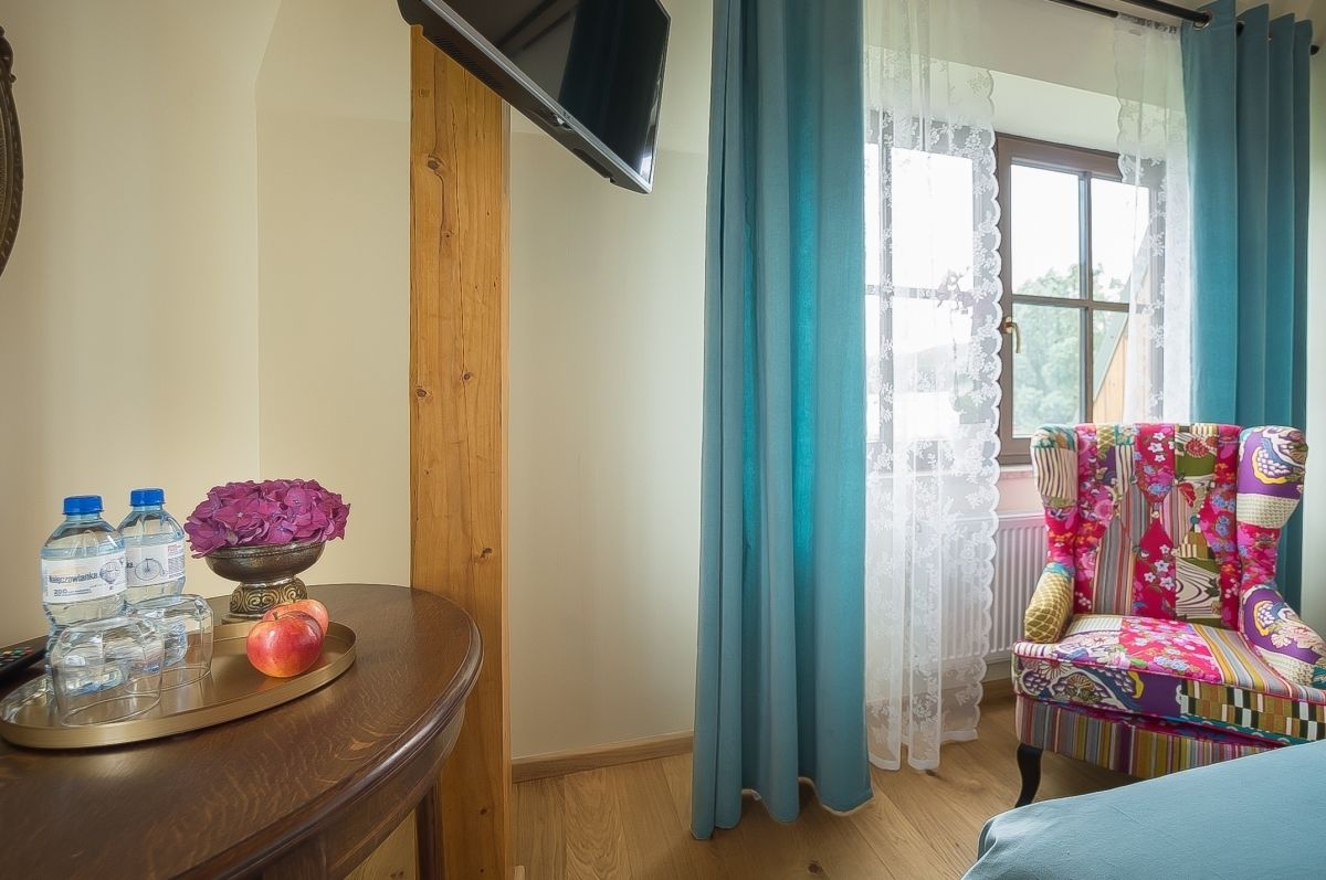 Una habitación con un escritorio de madera, un televisor en la pared, una silla colorida y una ventana con cortinas azules.