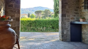 Une pièce aux murs de pierre, avec un plafond en bois rustique, une grande plante en pot et une vue sur un paysage verdoyant, avec des arbres et une colline au loin.