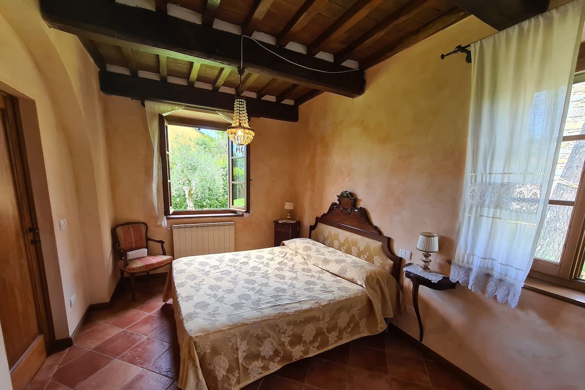 Un dormitorio con cama, una silla, una mesita de noche, un ventanal con cortinas y una lámpara colgante.