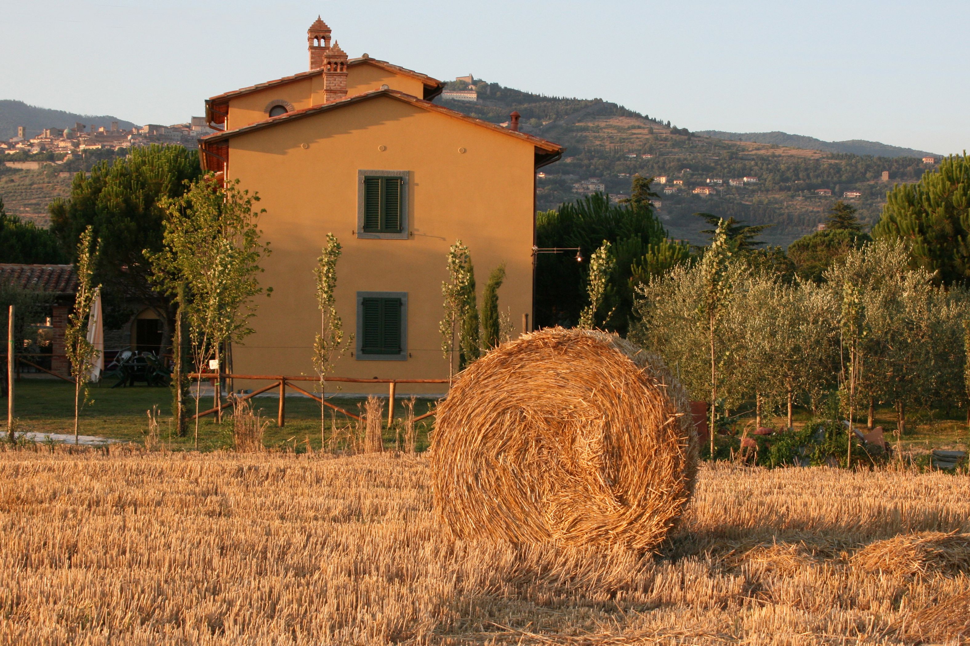 A rural house with a large hay bale in the foreground, surrounded by a field.