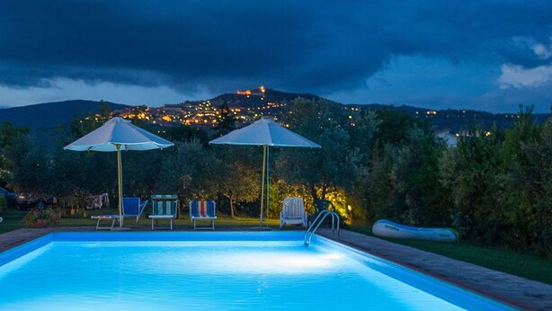 Une piscine avec deux parasols, entourée d’arbres, et offrant une vue sur une ville au crépuscule.