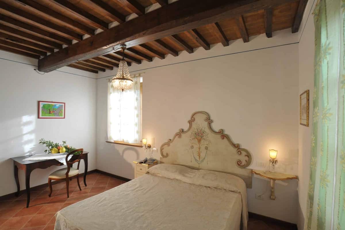 Un dormitorio con techo de madera, cama con cabecera ornamentada, escritorio con silla y ventana con cortina.