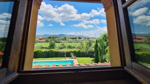 Une vue à travers une fenêtre ornée d’une arche en briques, donnant sur un paysage verdoyant avec une piscine, des arbres et des collines lointaines.