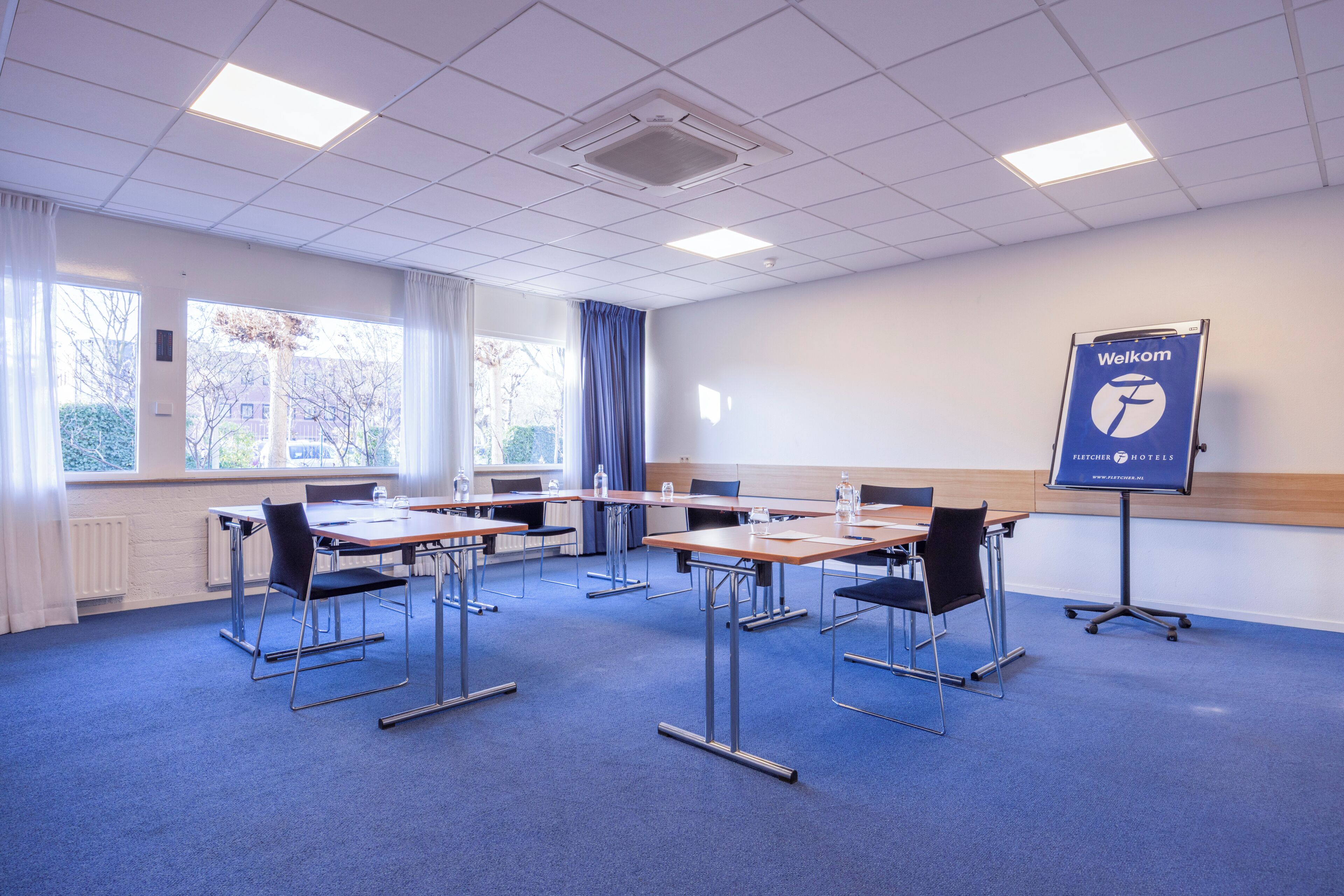 Una sala de conferencias con mesas y sillas, una alfombra azul y un cartel de 'Welcome'.