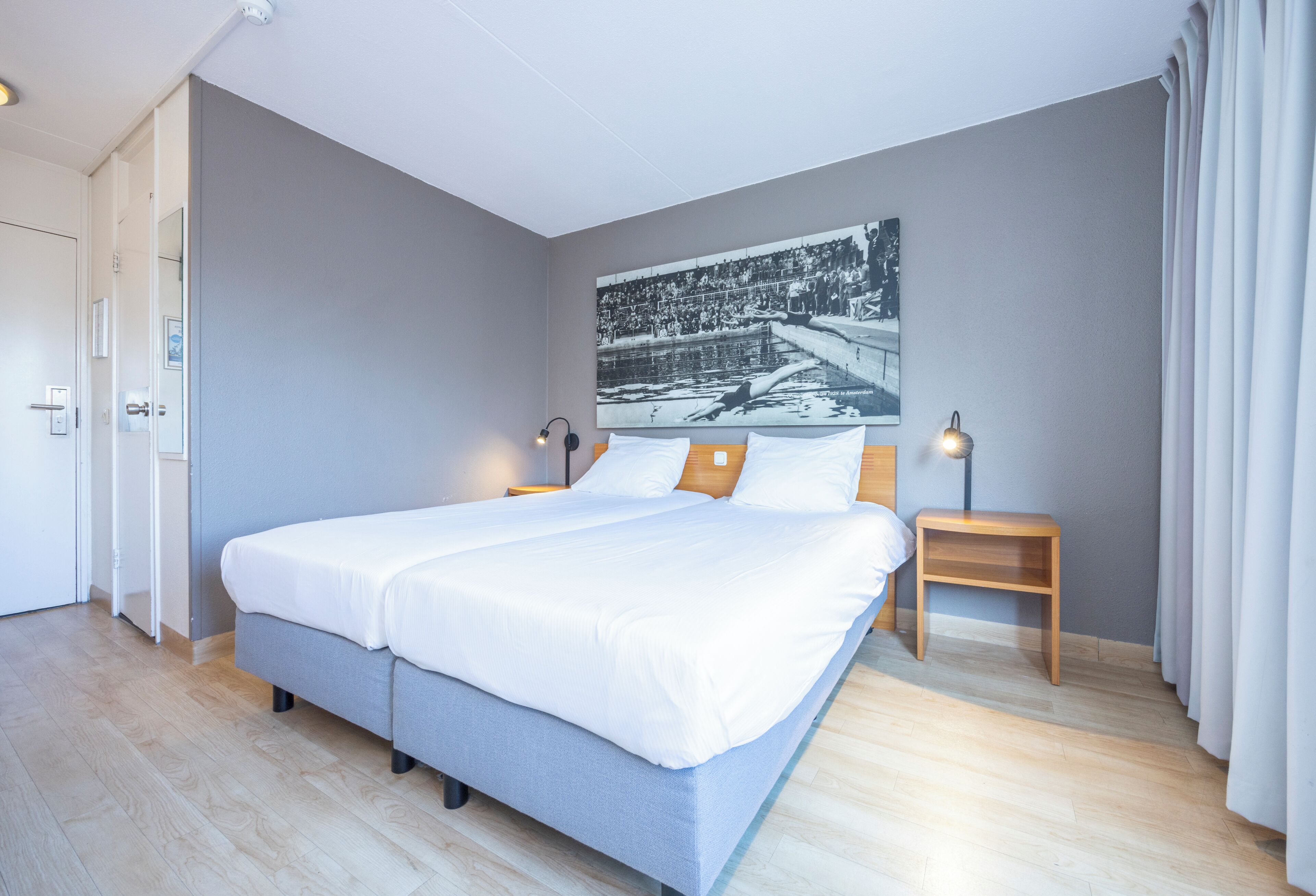 Quarto de hotel moderno com uma cama grande, mesa de cabeceira, abajur fixado na parede e uma porta.