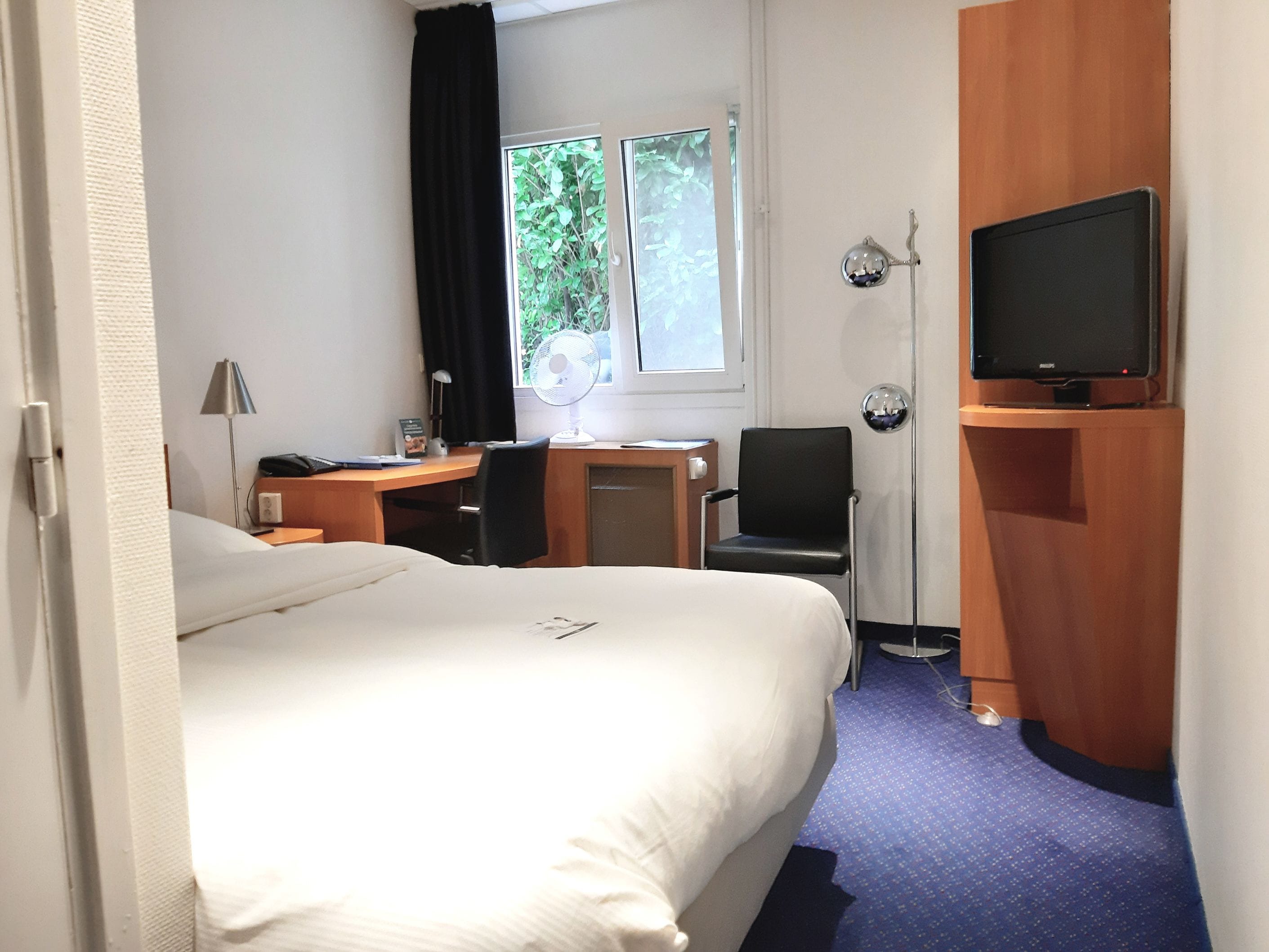 Habitación de hotel con cama, escritorio, silla, televisor y ventana con vista a follaje.