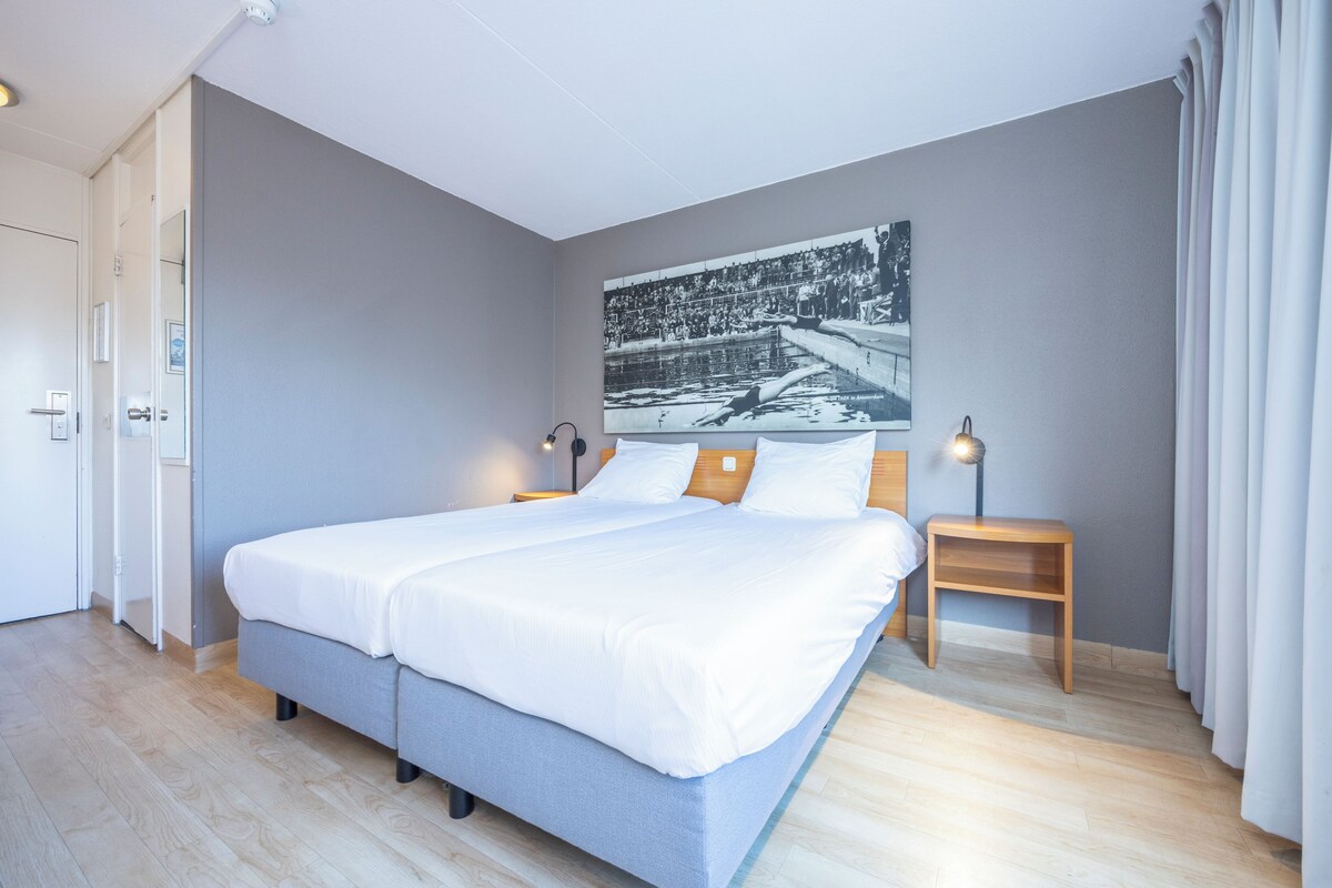 A modern hotel room with a large bed, a nightstand, a wall-mounted lamp, and a door.