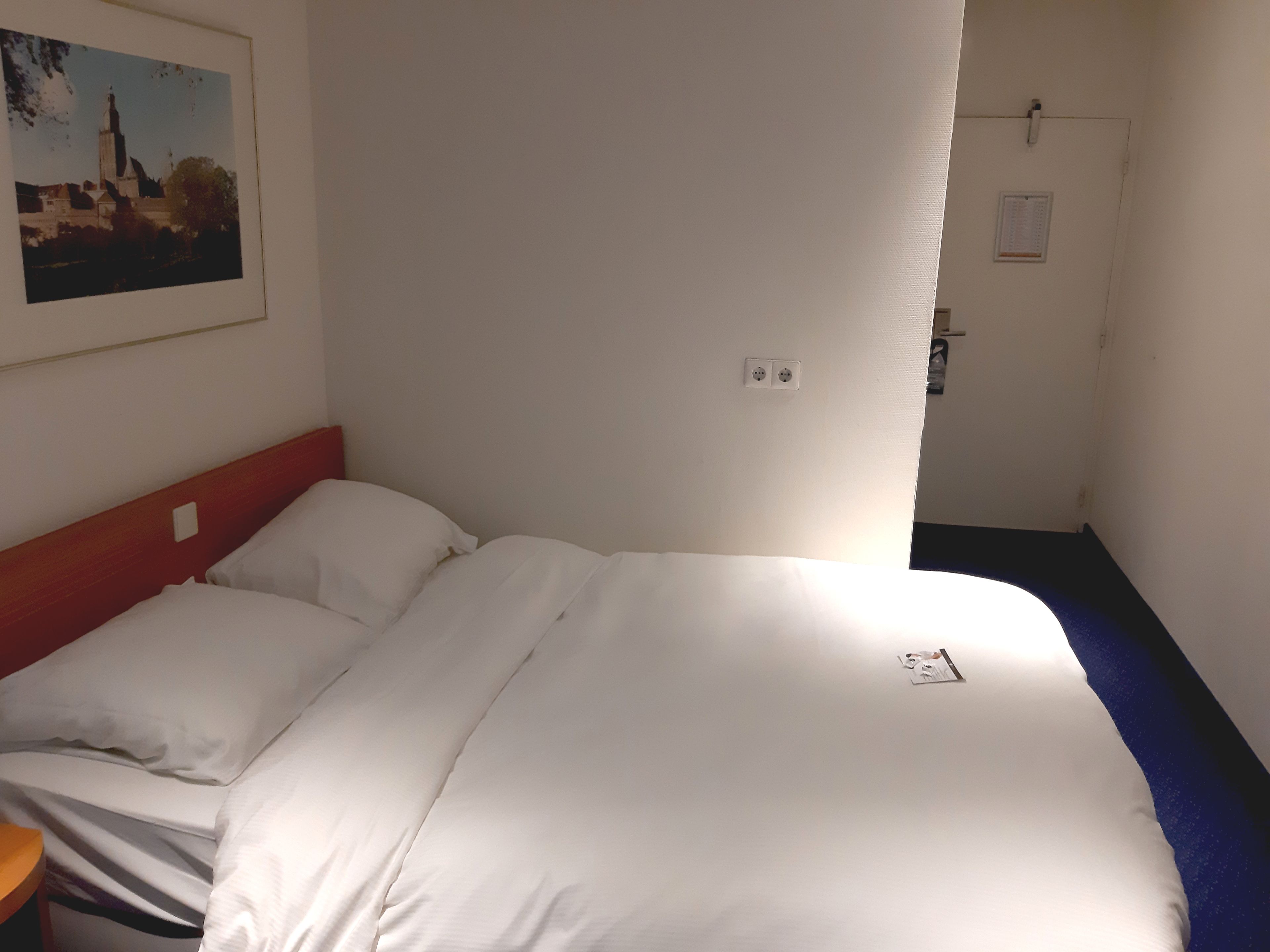 A hotel room with a bed, a nightstand, a painting on the wall, and a door with a light switch.