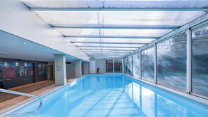 Una piscina interior con paredes de vidrio, una terraza de madera y vista a árboles.