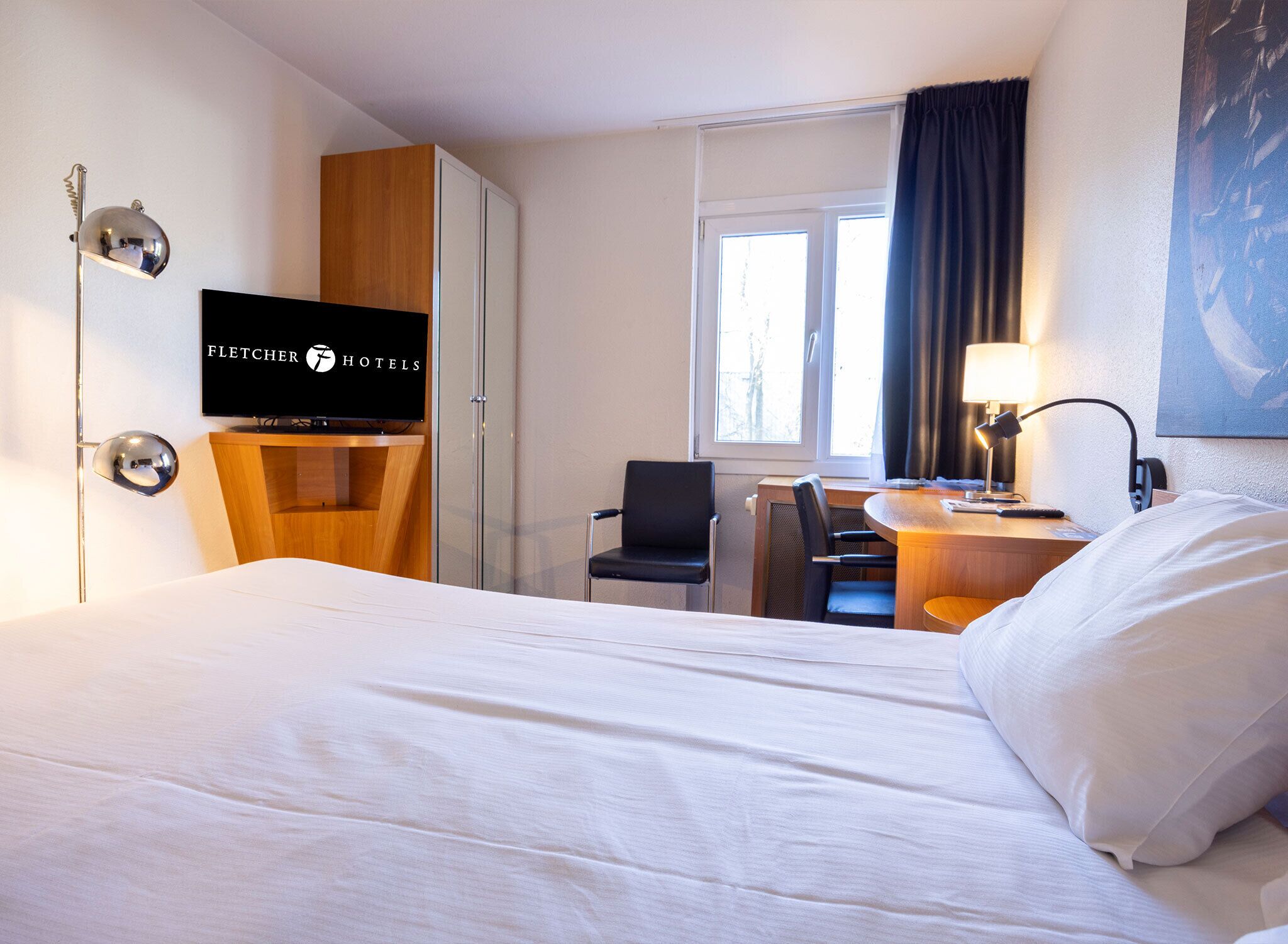 A hotel room with a bed, a desk with a chair, a television, and a window with curtains.