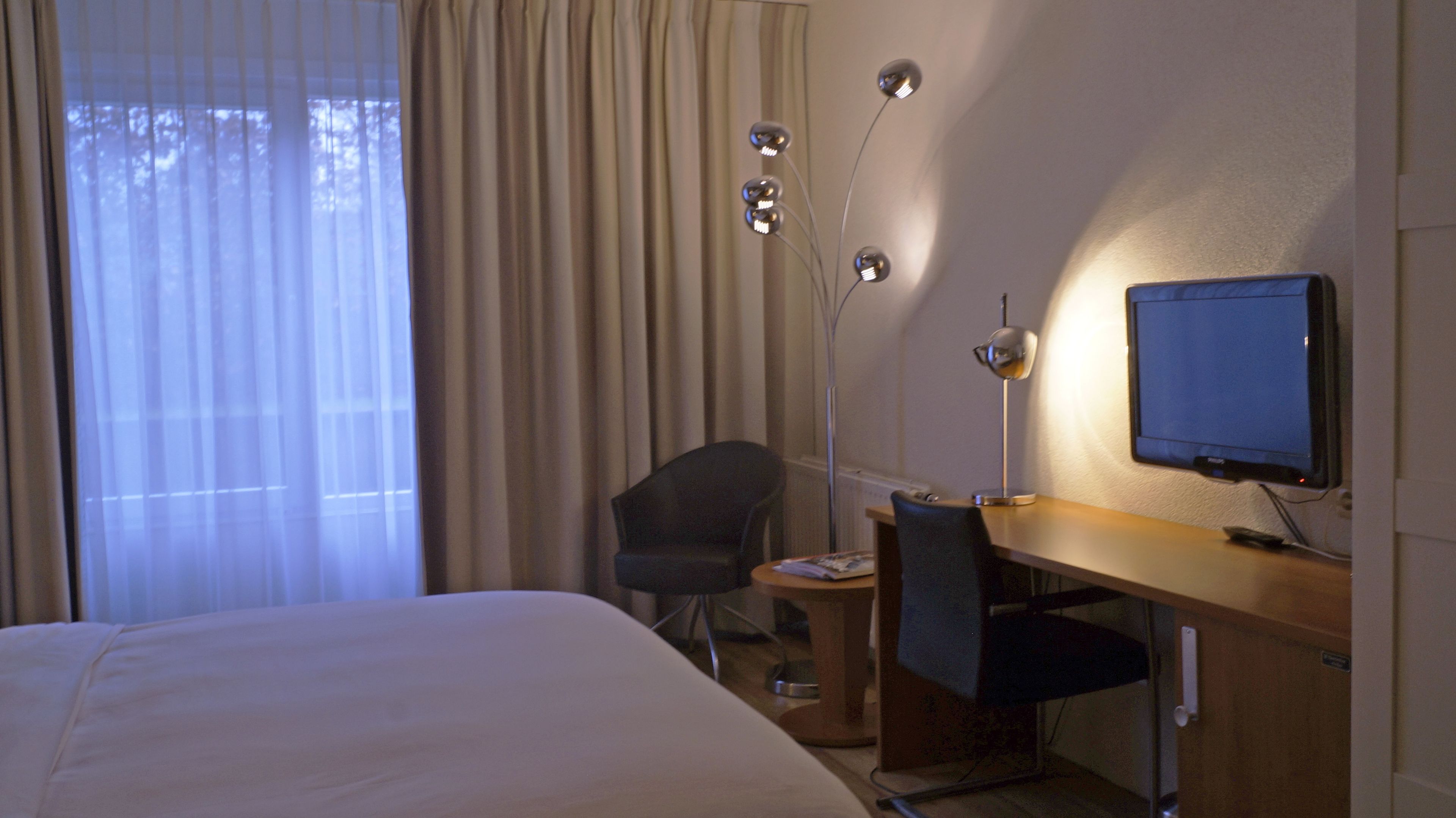 Ett hotellrum med en säng, ett skrivbord med en stol, en tv och en golvlampa.