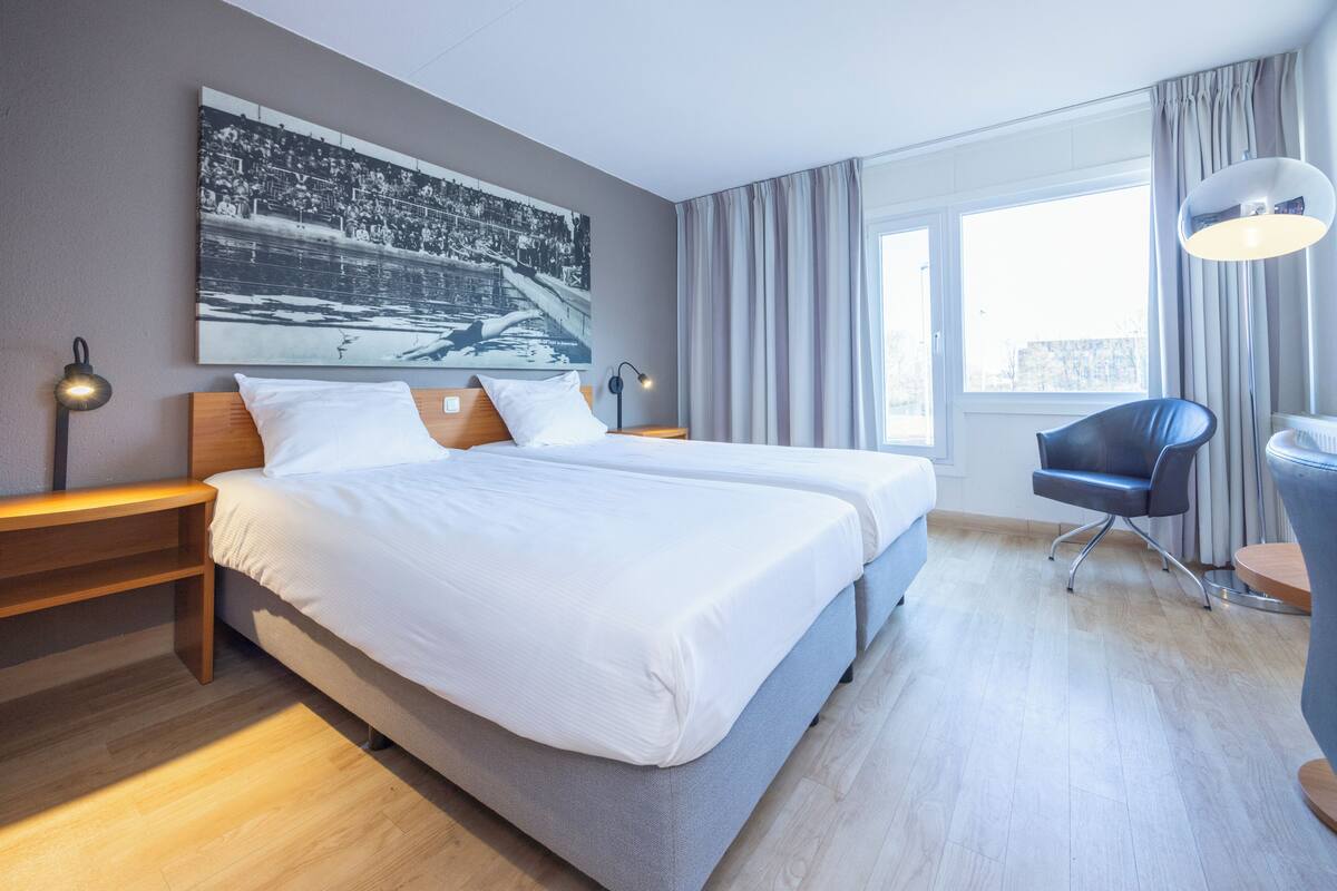 Habitación de hotel con una cama grande, dos mesitas de noche, una silla, un escritorio, una lámpara y un ventanal con cortinas.