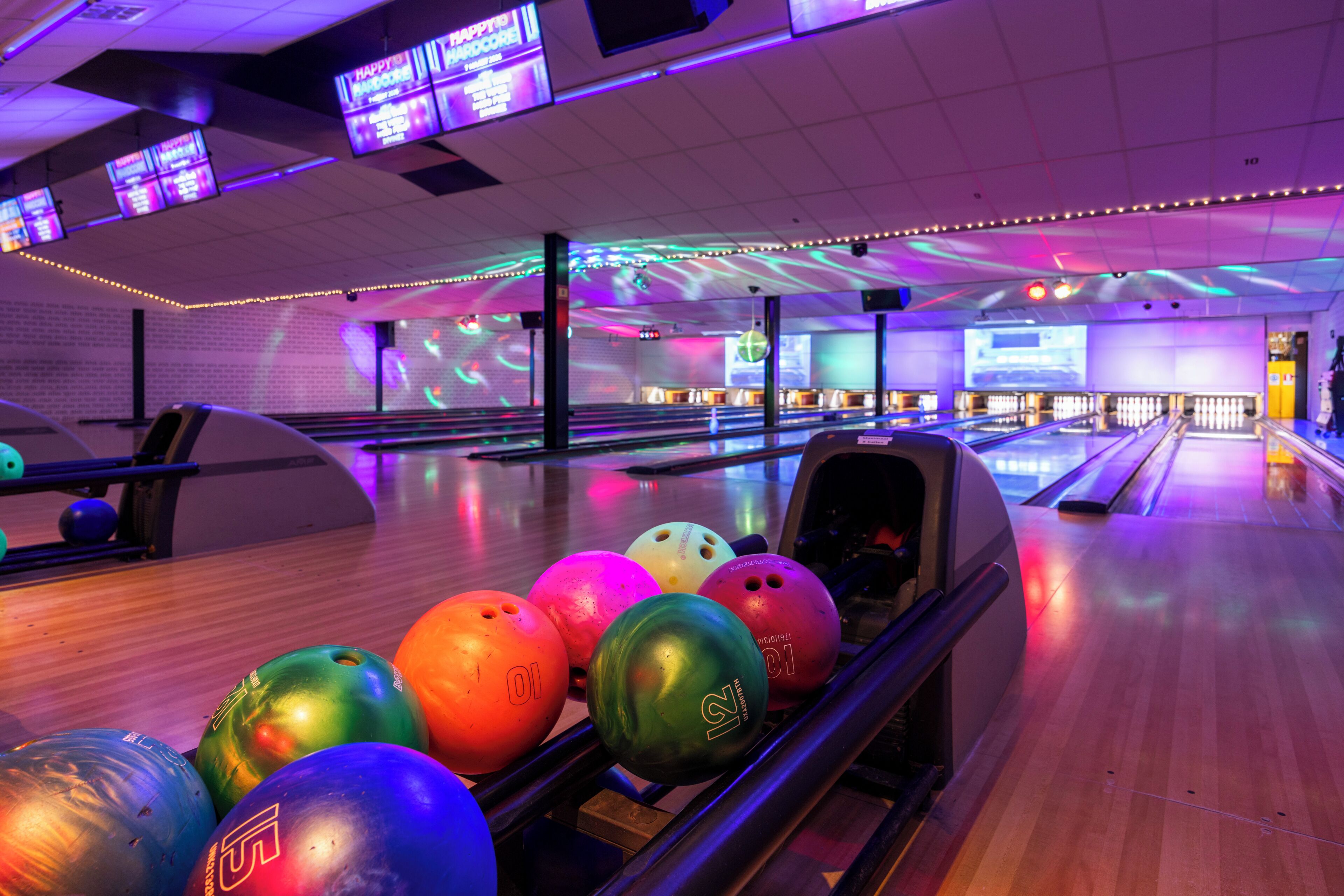 A bowling alley with multiple lanes, bowling balls, and overhead screens displaying advertisements.