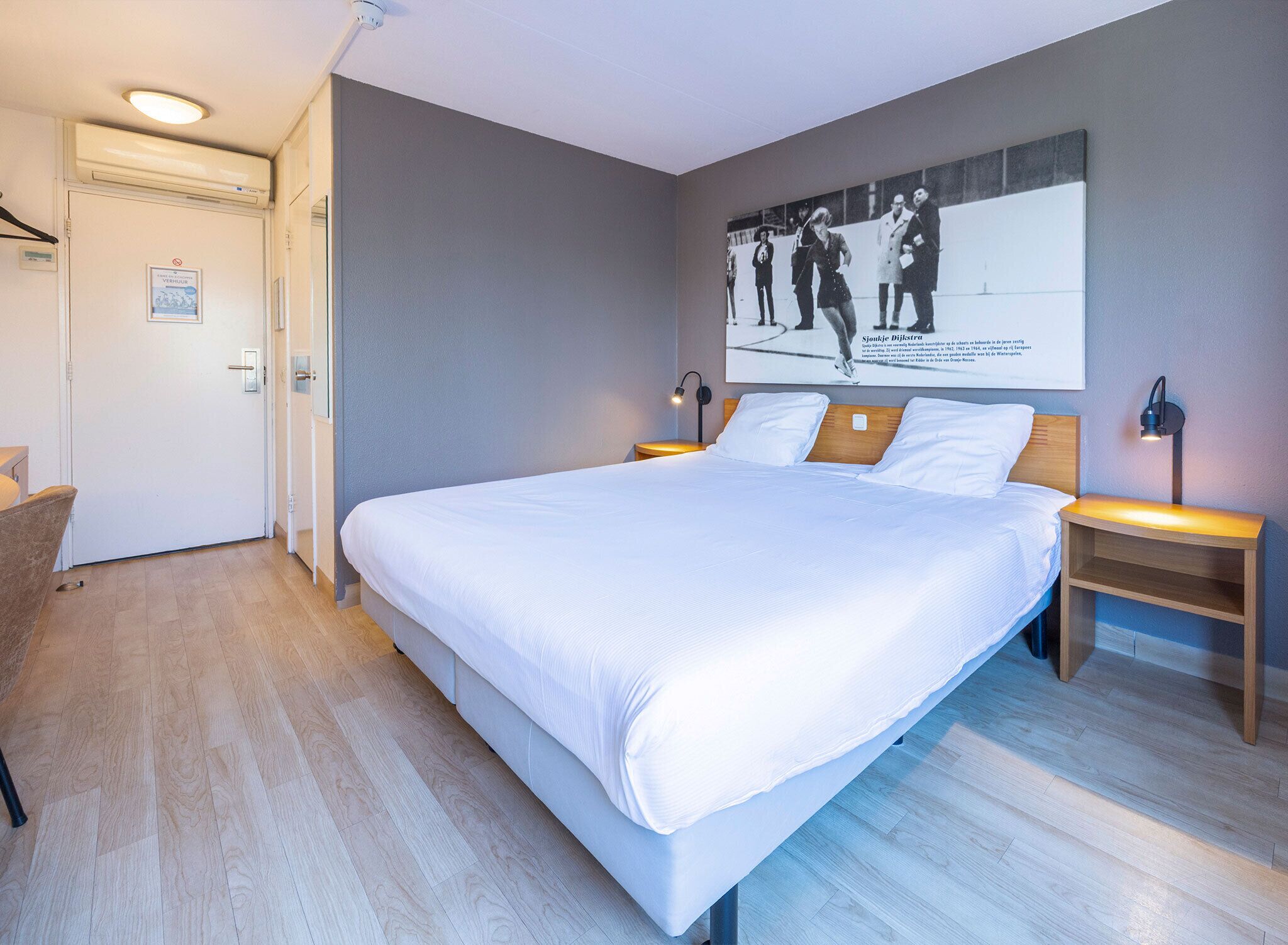 Una habitación de hotel con una cama grande, dos mesitas de noche, un cuadro colgado en la pared y una puerta con un cartel.
