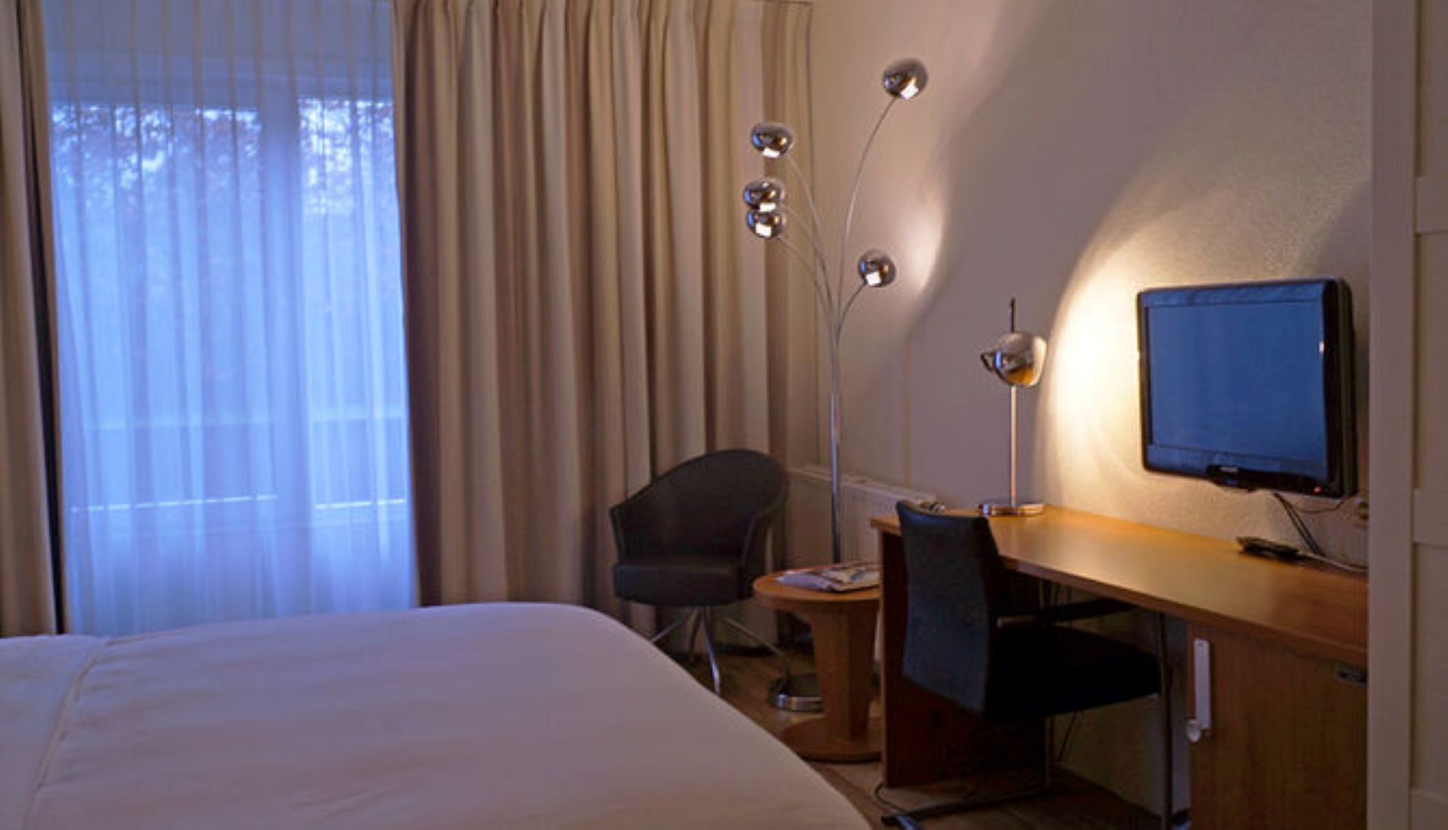 A hotel room with a bed, a desk, a chair, a desk lamp, and a television.