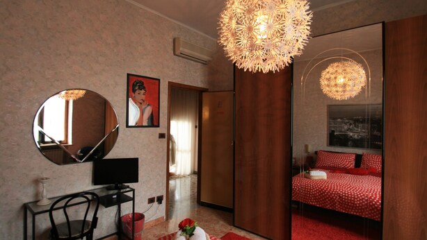 Een slaapkamer met een grote spiegel, een bureau met een computer, een rood stippen vloerkleed en een bed met rode beddengoed.