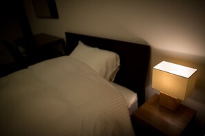 A neatly made bed with a white comforter and pillows, a bedside table with a lamp, and a dark headboard. - Rota Hostel (Osaka)