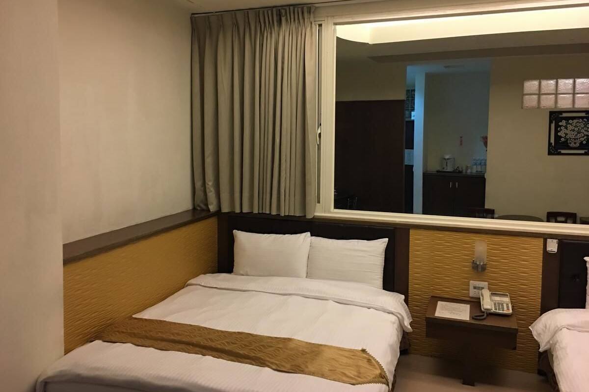 A hotel room with a bed, a bedside table with a phone and a lamp, a window with curtains, and a mirror reflecting the room.