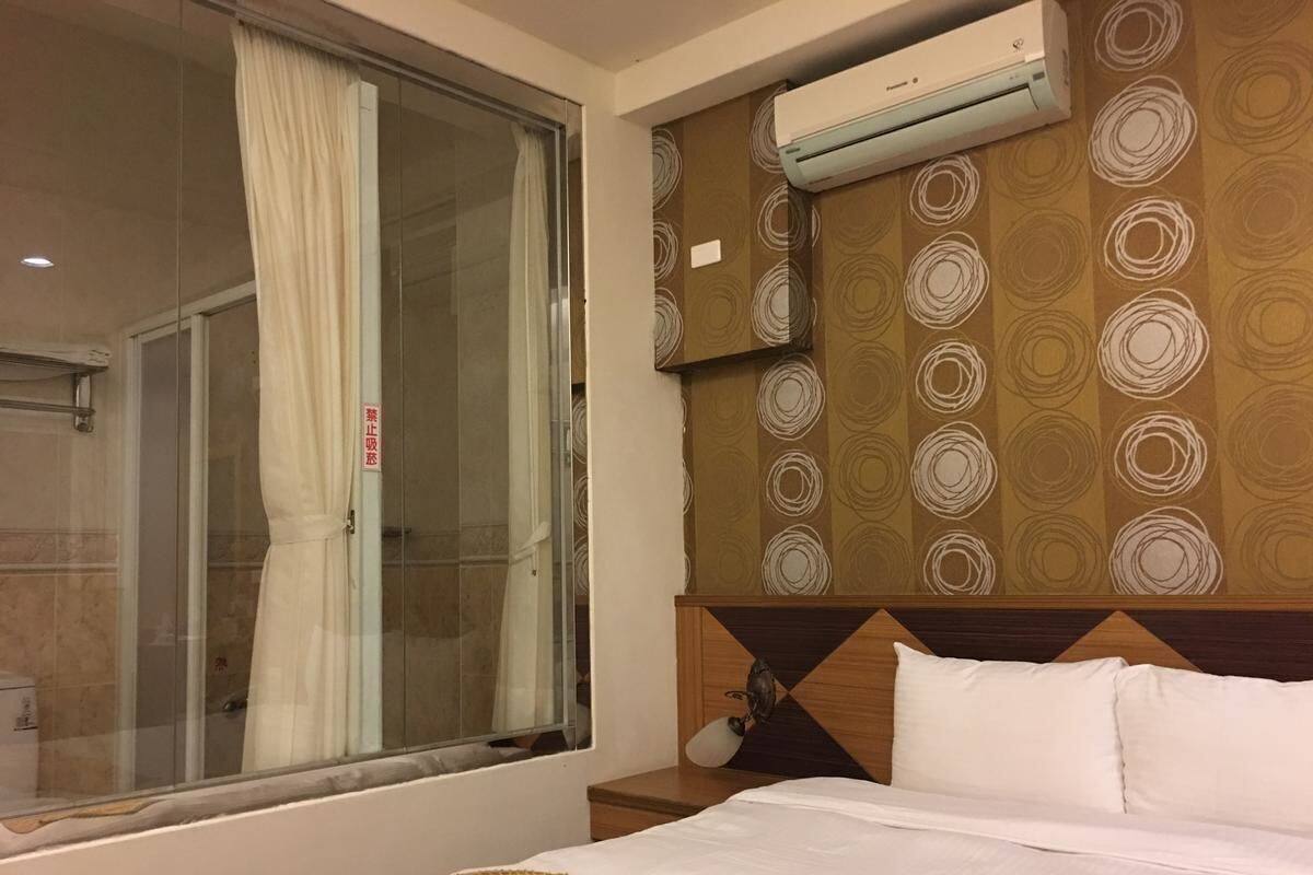 A hotel room with a bed, a window with curtains, a shower, and an air conditioning unit.
