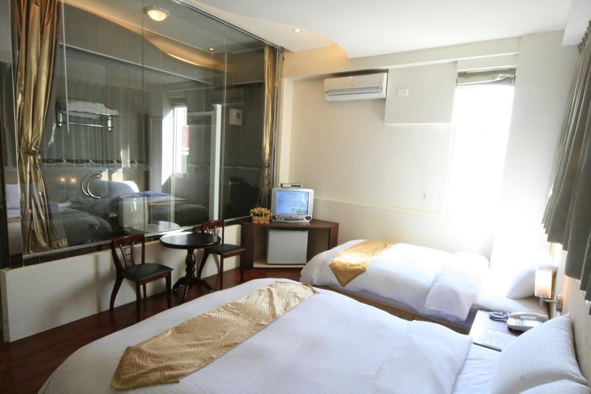 A hotel room with two beds, a desk with a computer, a chair, and a window with curtains.