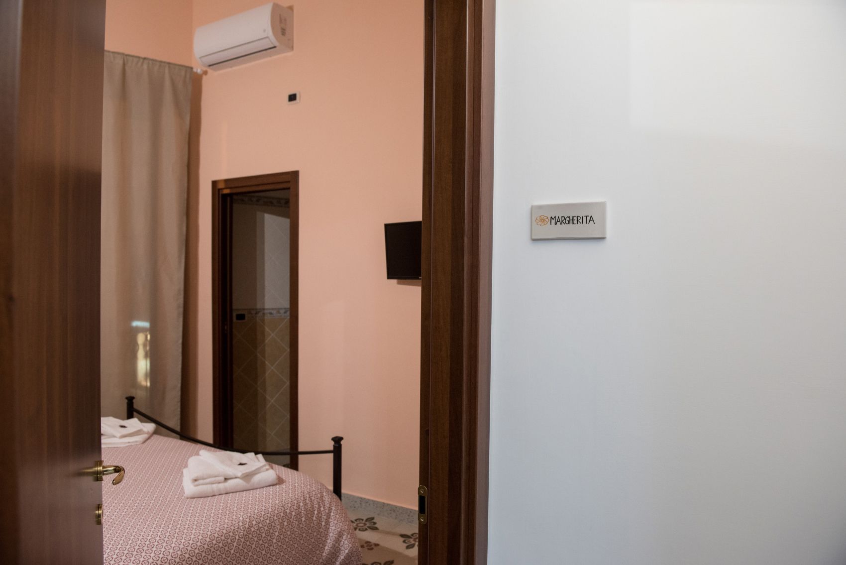 A bedroom with a bed, a door, a wall-mounted air conditioner, and a sign reading "MARGHERITA".