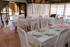 Un comedor con mesas y sillas blancas, techo de paja y cortinas.