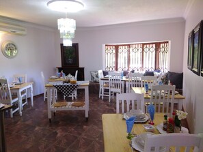 A dining area with white chairs and tables, a chandelier, and a window with decorative bars. - Tuscany Boutique Hotel (Vryburg)