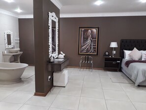 A modern bedroom with a large bed, a nightstand, a side table, a chair, a painting, and a bathtub. - Tuscany Boutique Hotel (Vryburg)