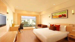 A hotel room with a large bed, a desk with a chair, a TV, a painting on the wall, and a window with a city view. - Lemon Tree Hotel Banjara Hills (Hyderabad)