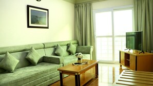 A living room with a green sofa, a wooden coffee table, a television on a wooden cabinet, and a large window with curtains. - Al Nile Hotel (Salalah)