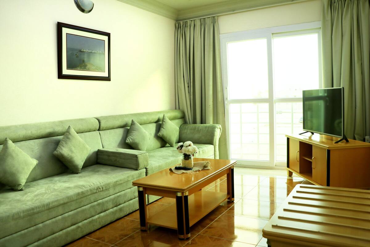 A living room with a green sofa, a wooden coffee table, a television on a wooden cabinet, and a large window with curtains.