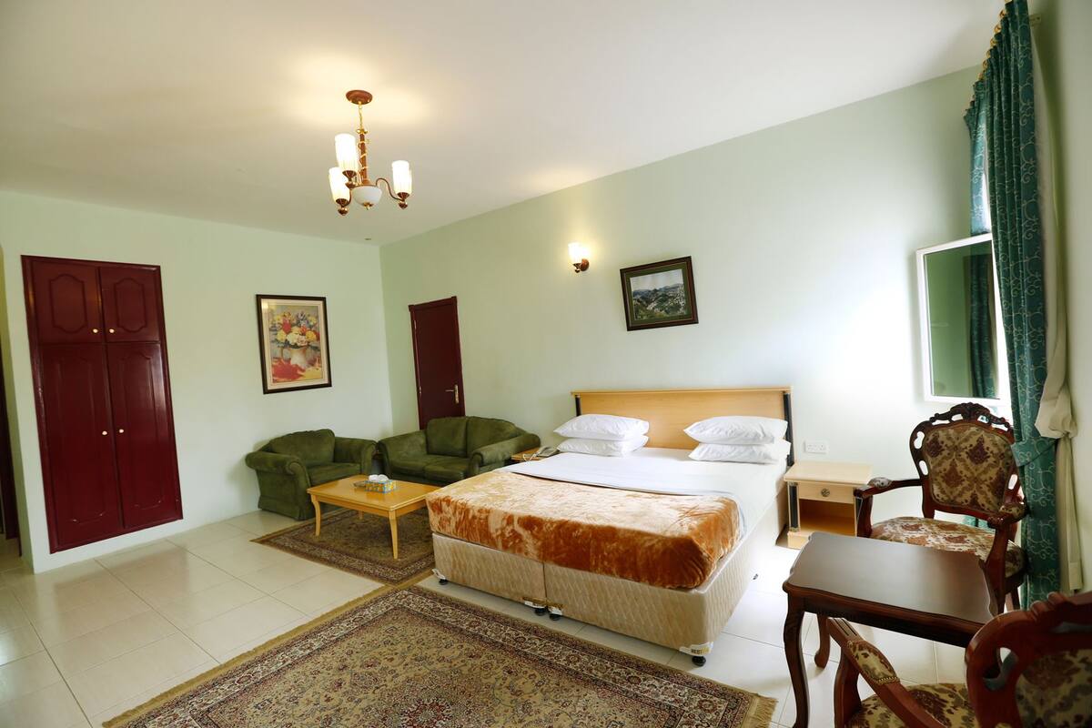 A hotel room with a bed, two green armchairs, a wooden table, a desk, and a chair.